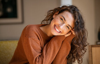 Smiling woman with curly hair, wearing a brown sweater, rests her head on her hands while seated indoors.