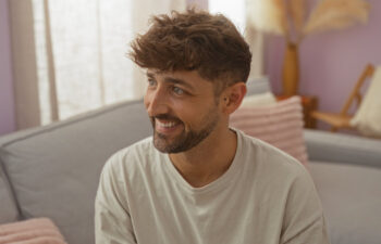 A man with short, wavy brown hair and a beard smiles while sitting on a light-colored couch in a softly lit living room.