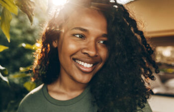 A person with curly hair and a green shirt smiles outdoors, sunlight shining through trees in the background.