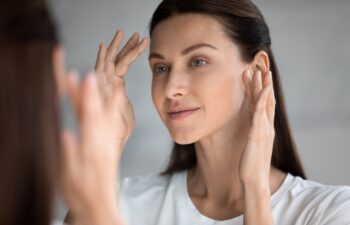 Close up beautiful woman looking in mirror in bathroom, touching face, pretty young female checking makeup, satisfied by healthy perfect skin, natural beauty concept, face care and treatment